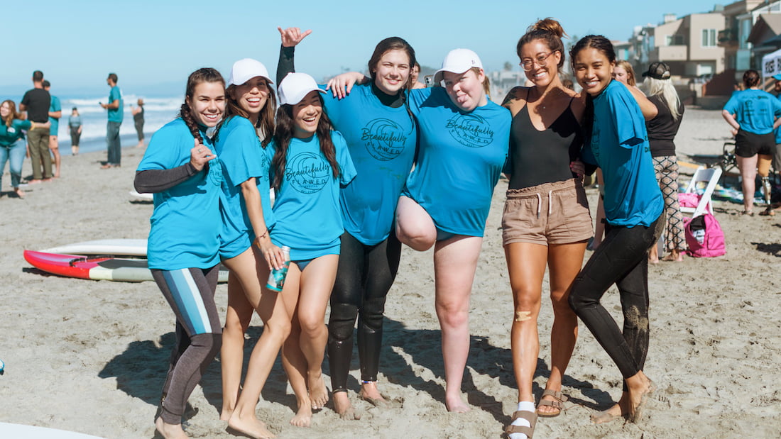 A group photo of seven Beautifully Flawed attendees and staff laughing and smiling on the beach during surf day!
