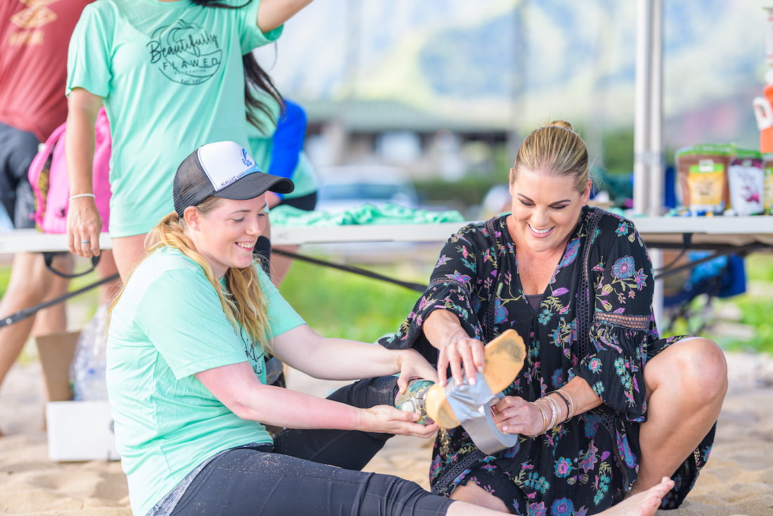 Sarah Hill, Beautifully Flawed Foundation Executive Director, helps a retreat attendee put tape on her prosthetic leg before going into the ocean to surf on surf day!