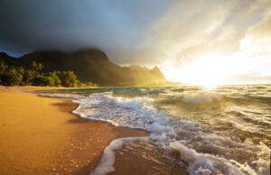 Golden hour photo at Tunnels Beach in Kauai with turquoise blue and white foam ocean waves crash on the sand with Kauai's green mountain range in the near distance.