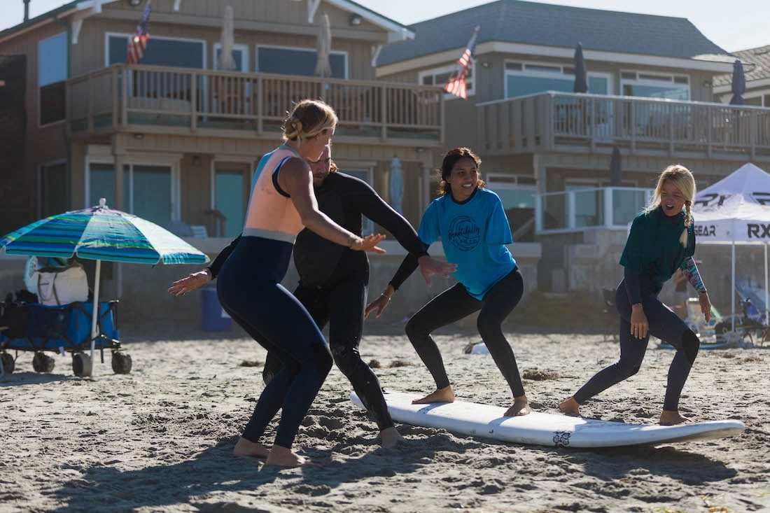 Bethany Hamilton and other surf instructors showing Beautifully Flawed attendee Ellice on the sand how to properly stand and balance on a surfboard as well as how to adapt with her arm limb difference.