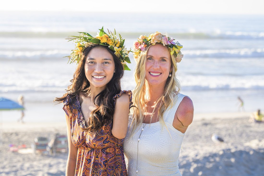 A beautiful photo of Bethany Hamilton and Ellice with their haku leis smiling on the beach with waves crashing in the background as they show off their arm limb differences.