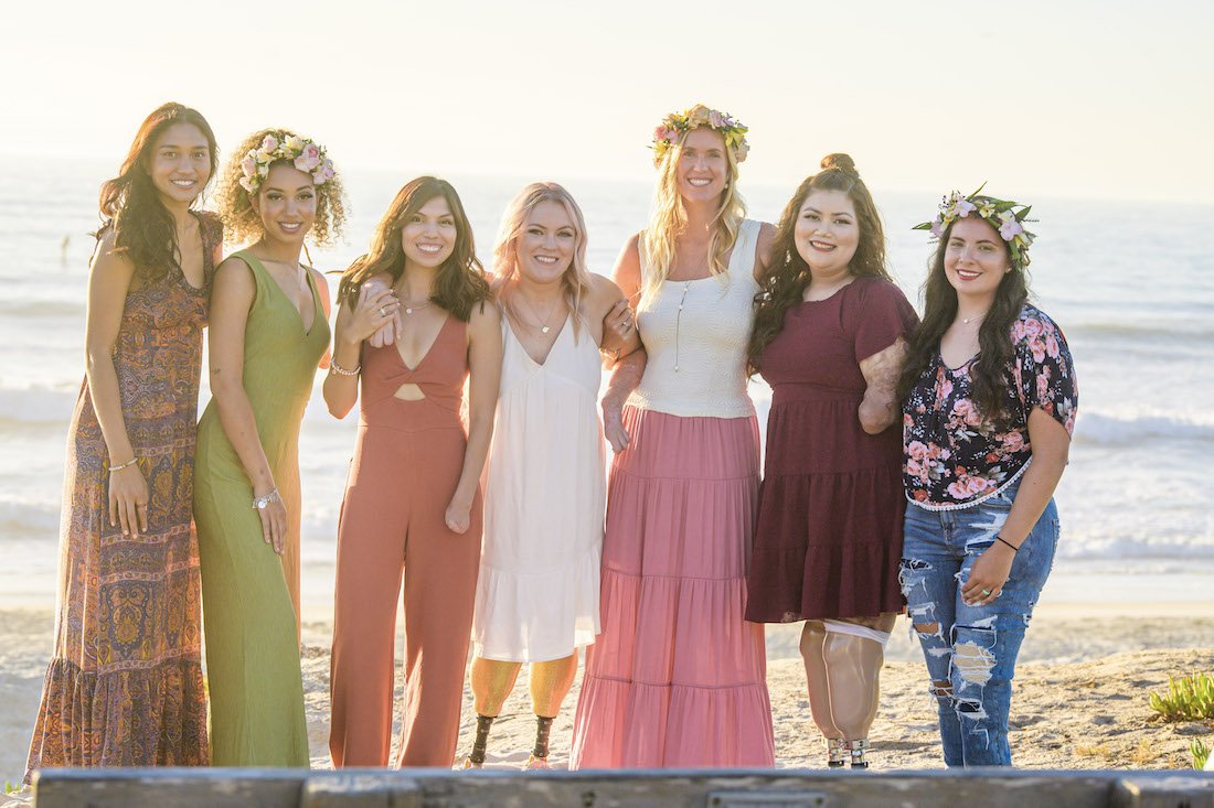 Ellice and five other young women amputees smile in a beach group photo during golden hour with Bethany Hamilton wearing haku leis and their prostheses.