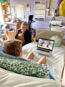 Ainsley sitting in her hospital bed watching Bethany Hamilton share in one of the Beautifully Flawed retreat recap videos and Sarah Hill watching with her.